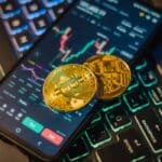 Cryptocurrency trading interface on a mobile with Bitcoin coins on a laptop keyboard.