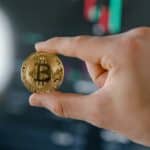 Close-up of a hand holding a Bitcoin against a stock market chart backdrop, symbolizing cryptocurrency investment.