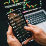 Hands holding smartphone with stock app, laptop displaying charts. Perfect for financial and technology themes.