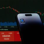 A mobile phone displaying a trading app in front of a financial market chart screen.
